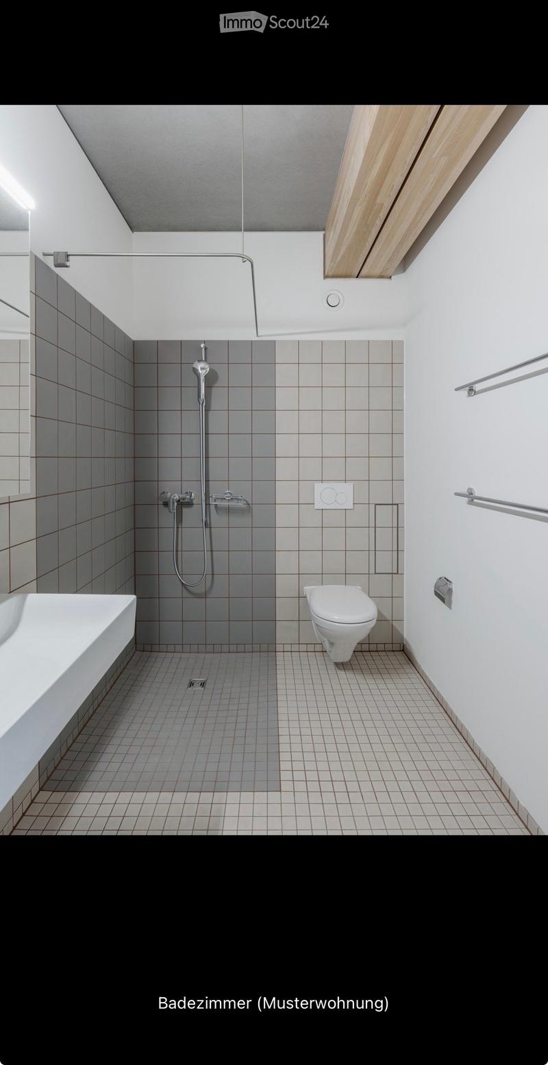Modern bathroom with a bathtub, toilet, and shower area. Features grey tiled walls and floor, sleek fixtures, and a wooden slatted ceiling.