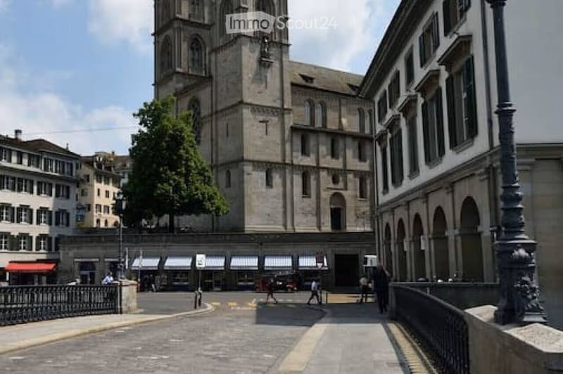 A large historic building with a tall tower, likely a cathedral, surrounded by smaller buildings and a paved walkway.
