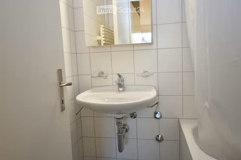 A small bathroom with a white sink, chrome faucet, and a mirror above it. The walls are tiled in light gray, and a door is visible on the left side.