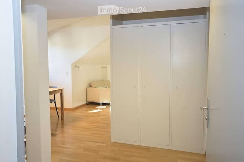 A small, well-lit room with wooden flooring, a built-in closet, a small table with a chair, and a lamp near the corner.
