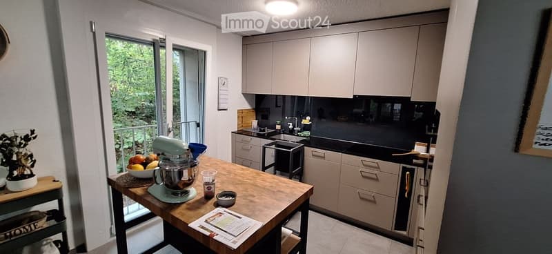 Modern kitchen with light grey cabinets, black countertops, and a wooden island. The island features a mixer, bowl, and other small items. A sliding glass door offers a view of greenery outside.