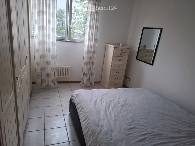 Bedroom with a single bed, dresser, mirror, and radiator. Light-colored walls and tiled floor.