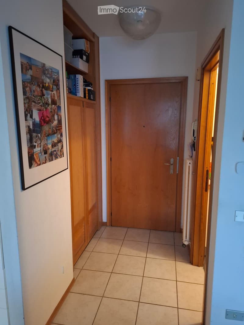 A well-lit hallway with tiled flooring, wooden doors, and a built-in wooden cabinet with shelves and a framed collage of photos on the wall.