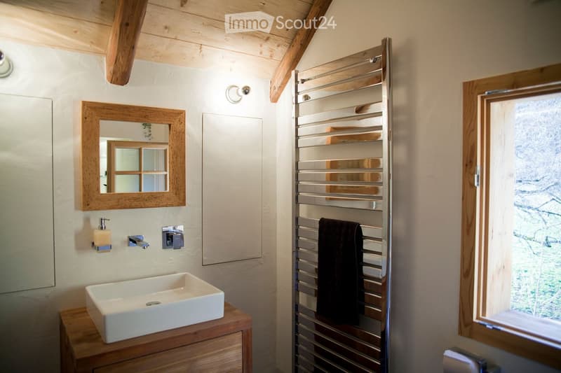 Bathroom with a white sink on a wooden vanity, a framed mirror, a towel rack with a dark towel, and a window showing greenery outside.