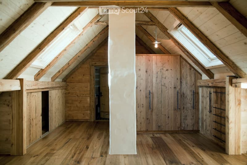An attic space with wooden beams, exposed wooden walls, and a large wooden wardrobe with multiple doors and drawers. The room features wooden flooring and skylights allowing natural light to enter.
