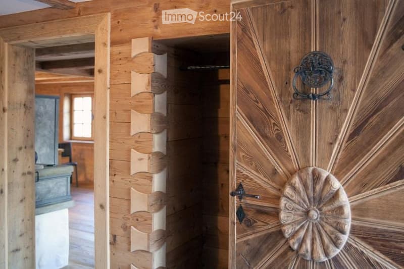 A wooden hallway with a decorative door featuring a sun-shaped handle and a carved design, leading to another room with visible wooden beams and a window.