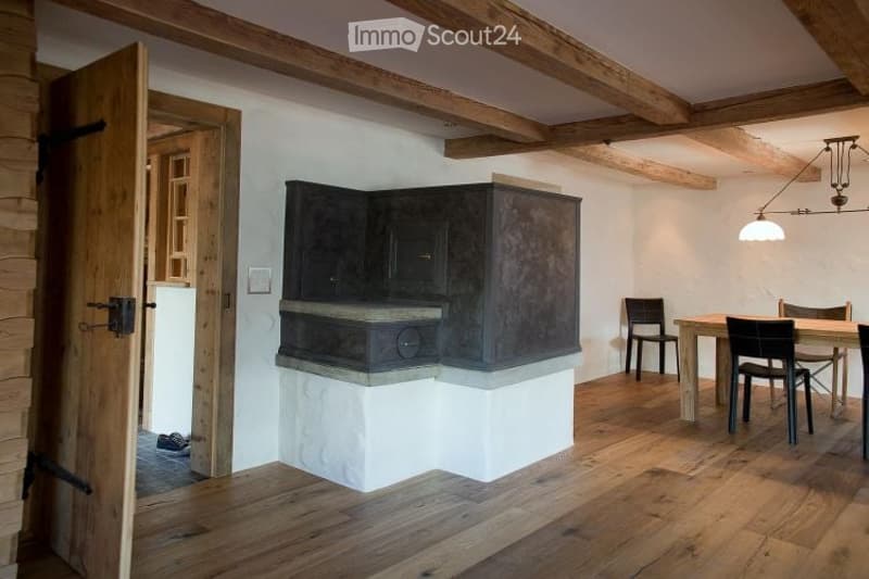A modern, rustic dining area with exposed wooden beams, a built-in stone dining counter, and a sleek black cabinet. The room features wooden flooring and a warm, inviting ambiance.