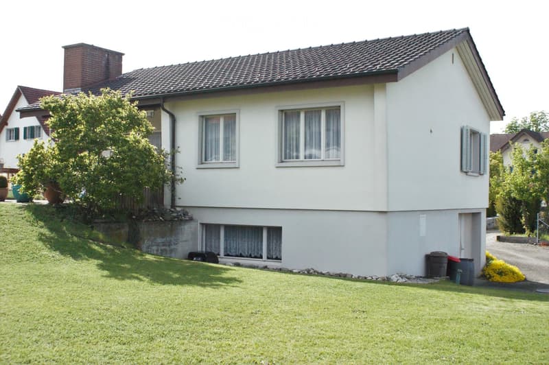 Two-story house with white exterior, tiled roof, and brick chimney. Features multiple windows with curtains, a raised ground floor, and a well-maintained lawn with trees and shrubs.