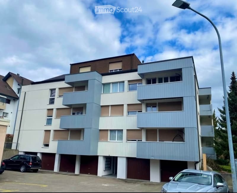 Modern multi-story apartment building with balconies, light blue and white facade, and parking area.