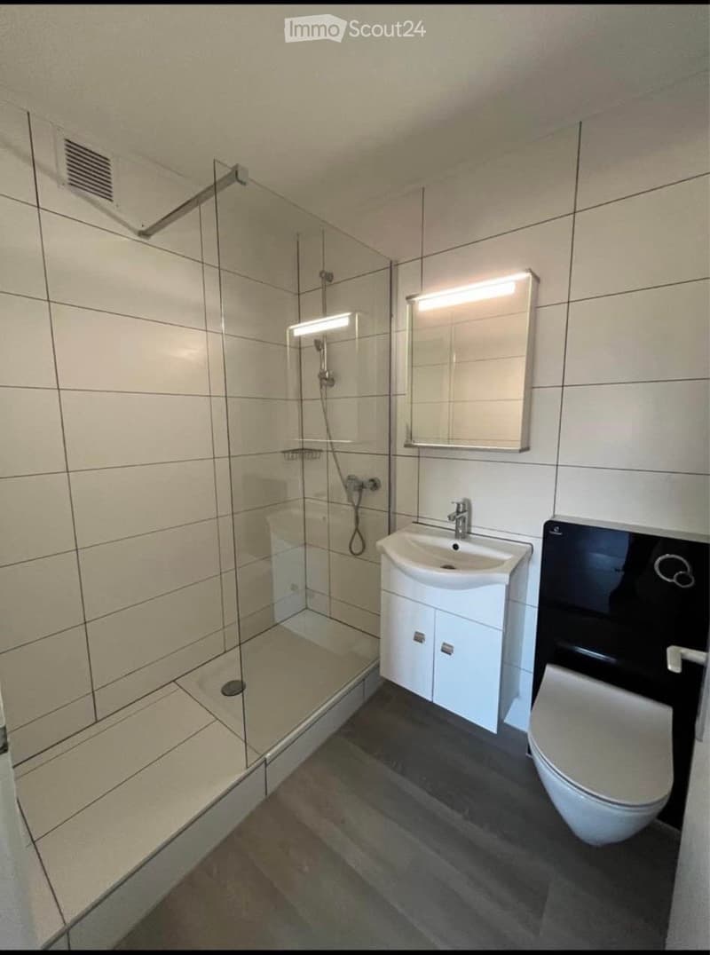 Modern bathroom with white tiles, a glass-enclosed shower, a white sink with a cabinet, a mirror with built-in lighting, and a toilet.