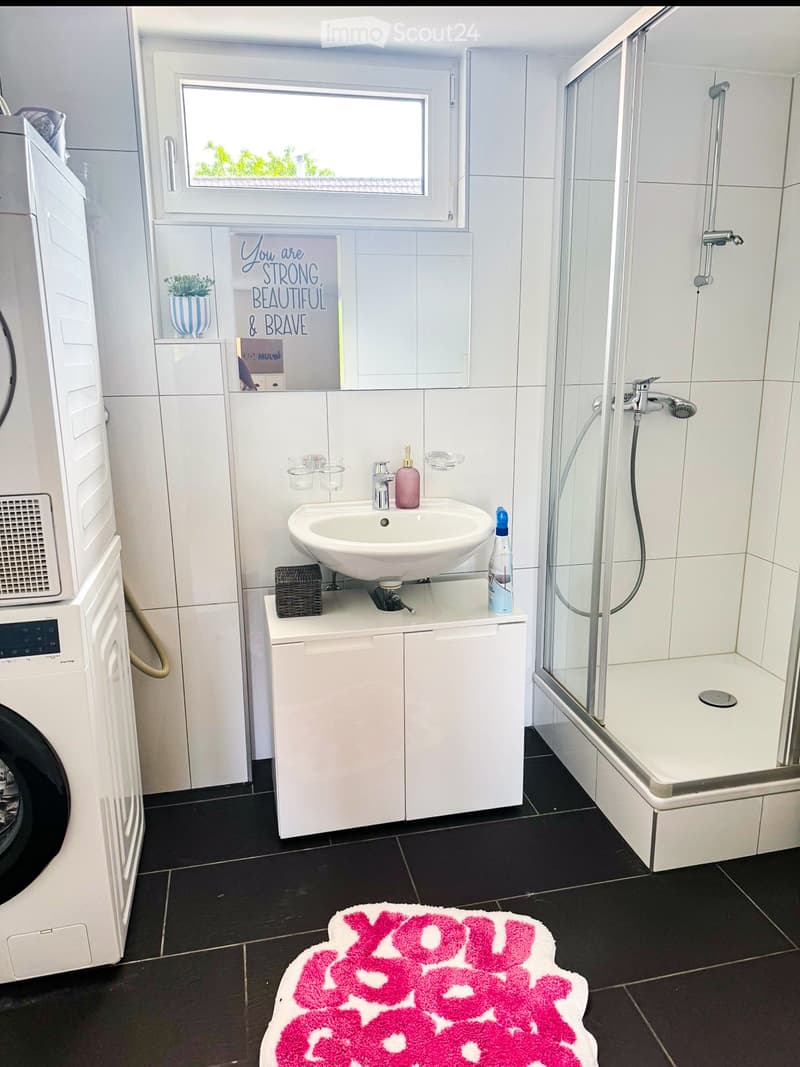 Modern bathroom with a white sink, shower, washing machine, and a decorative rug on the floor.