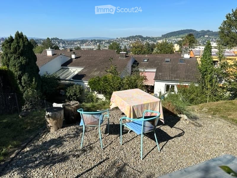 Outdoor terrace with a table and chairs, overlooking a scenic cityscape with houses and trees.