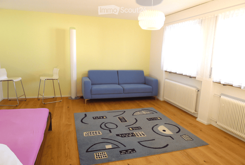 Bright living room with wooden flooring, blue sofa, two white stools, colorful rug, and a hanging lamp.
