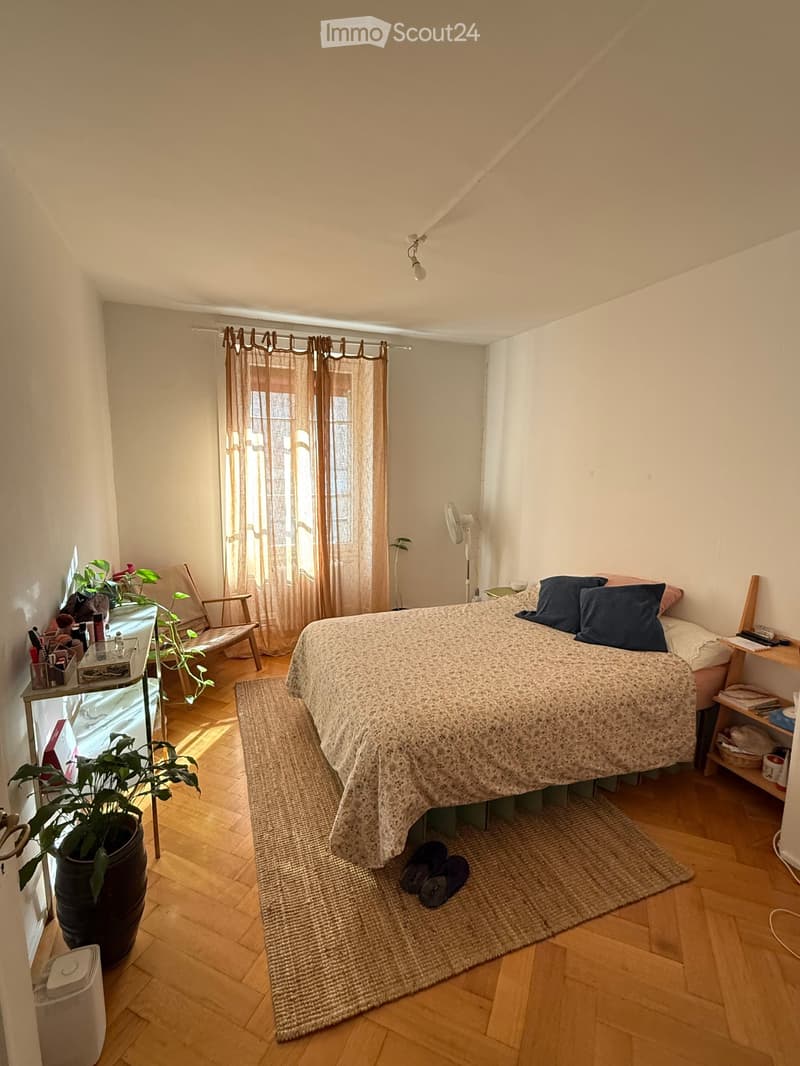 Cozy bedroom with wooden flooring, a bed with patterned bedding, a nightstand, a chair, a fan, a plant, and a small table with a mirror.