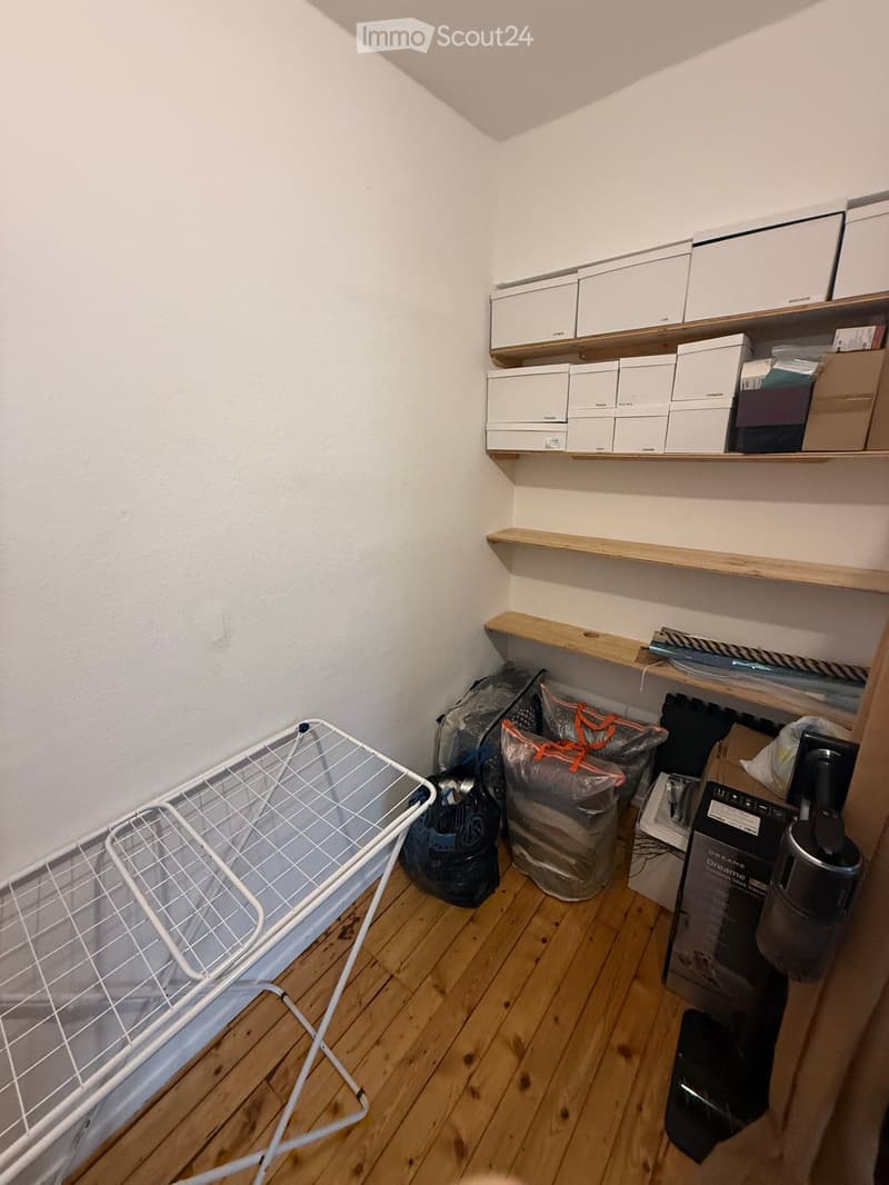 A small storage room with wooden shelves holding boxes and items, a laundry basket, and a vacuum cleaner on the floor.