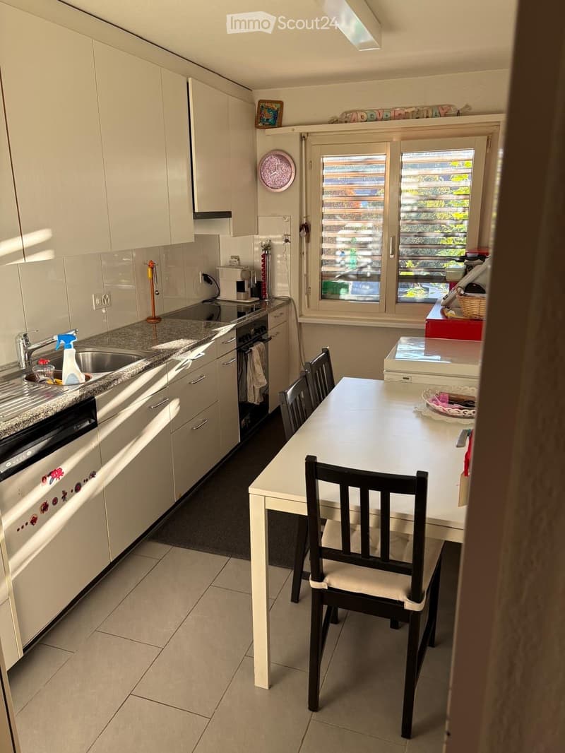 Modern kitchen with white cabinets, granite countertop, sink, oven, dishwasher, and a small dining area with a table and chair.