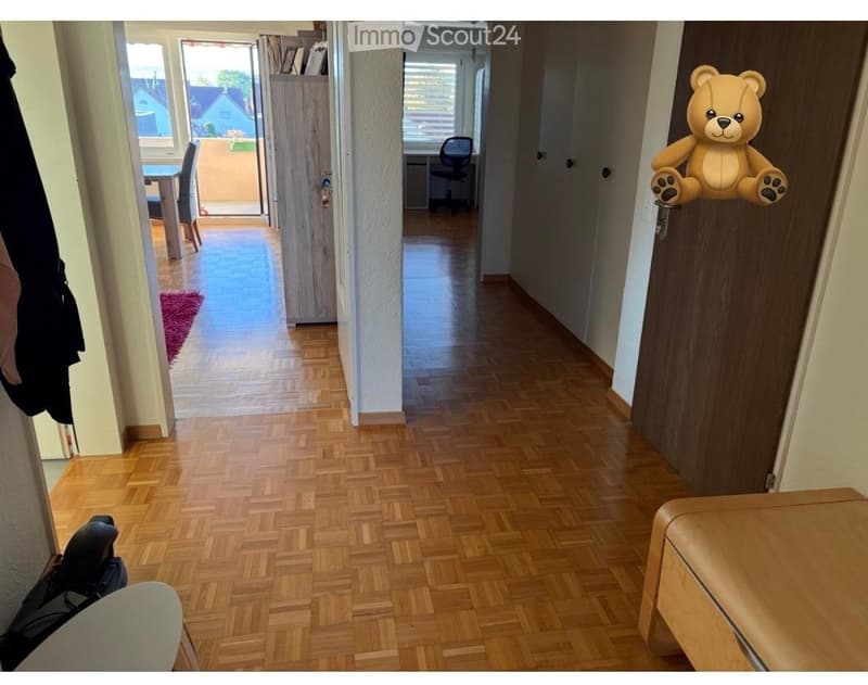Spacious hallway with wooden flooring, leading to a dining area and a room with a desk and chair. A large teddy bear is attached to the wall on the right.