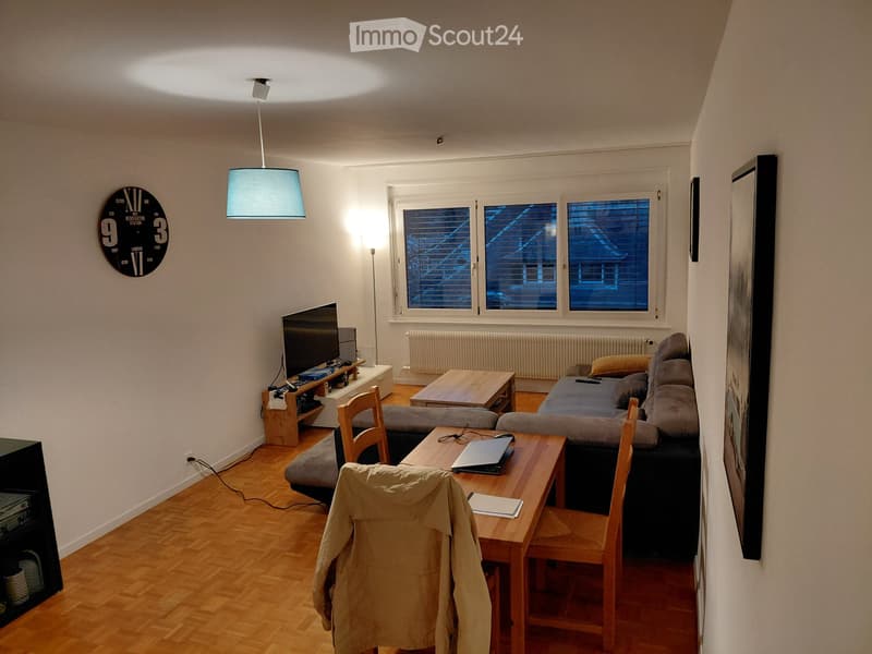 A modern living room with a grey sofa, coffee table, laptop on a wooden table, chairs, television, and a clock on the wall.