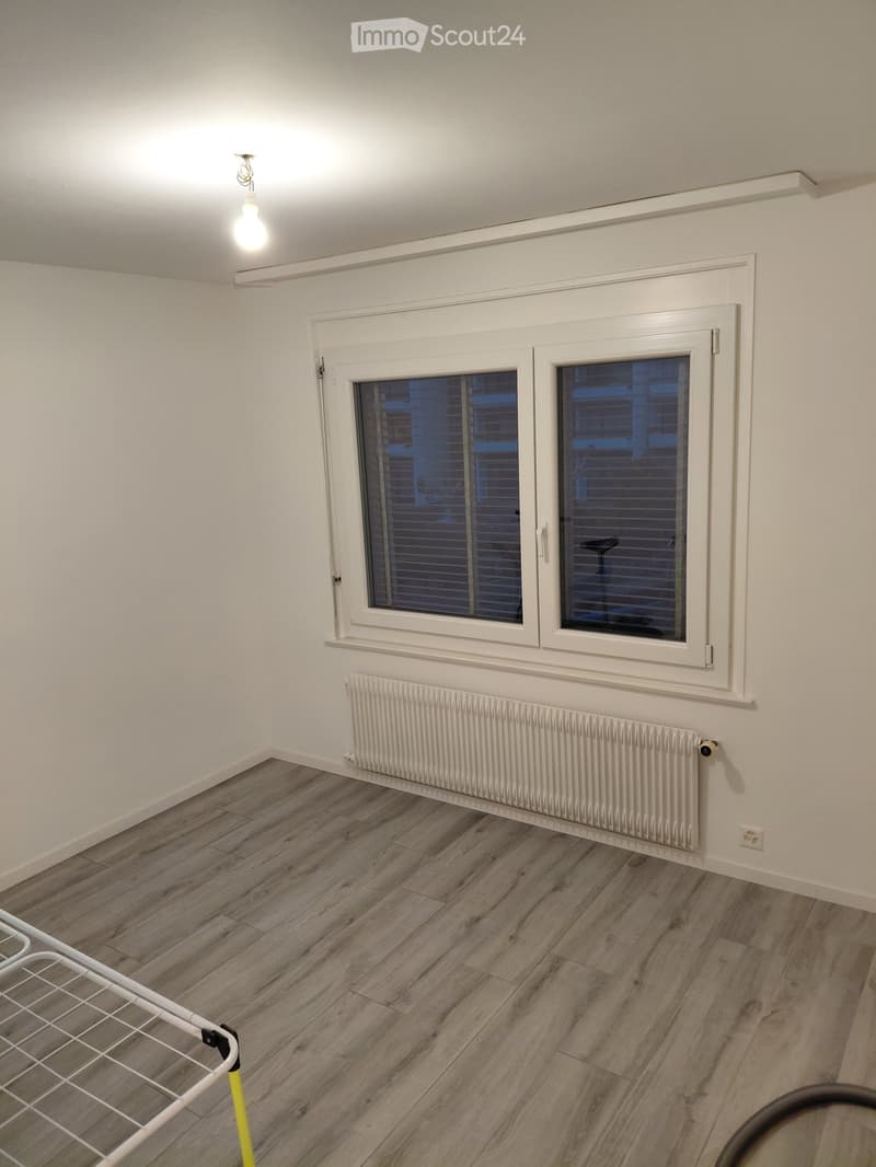 Empty room with wooden flooring, white walls, a radiator, and a window with blinds. A clothes drying rack is partially visible in the foreground.