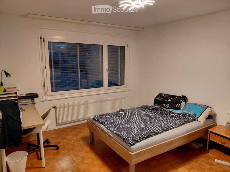 A tidy bedroom with a wooden bed, desk, chair, nightstand, and a window with blinds. The room features a wooden floor and white walls.