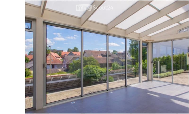 Bright, empty room with large glass walls and a skylight, offering a view of a suburban neighborhood.