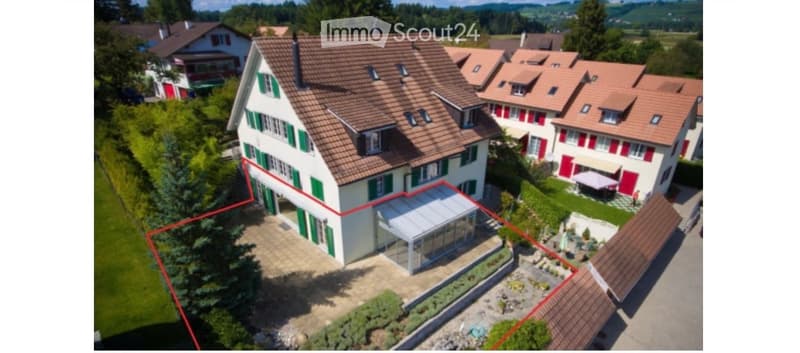 Aerial view of a two-story house with a red-tiled roof, green shutters, and a modern glass extension. The house is surrounded by a well-maintained garden and is part of a residential neighborhood with similar houses in the background.