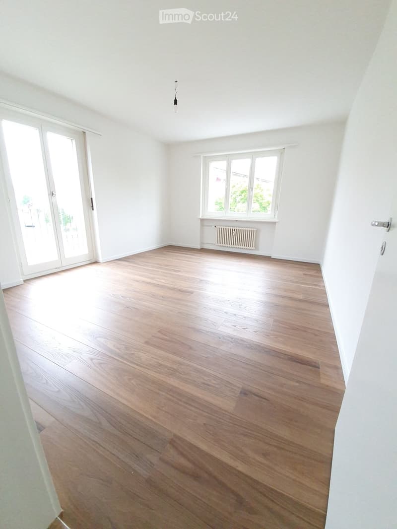 Empty room with light wooden flooring, white walls, large windows, and a sliding glass door leading outside.