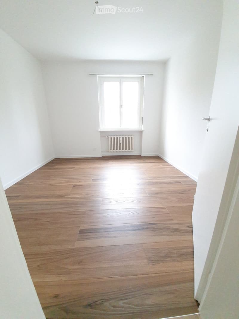 Empty room with wooden flooring, white walls, and a single window with a radiator below it.
