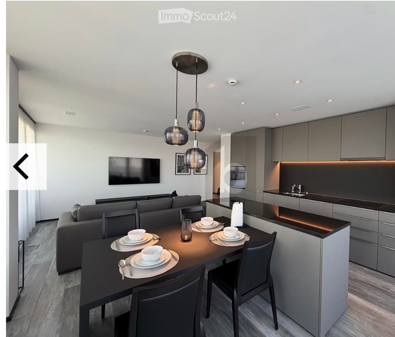 Modern open-plan living and dining area with a grey sofa, a black dining table with chairs, a sleek kitchen area with cabinets and a sink, and pendant lights hanging from the ceiling.