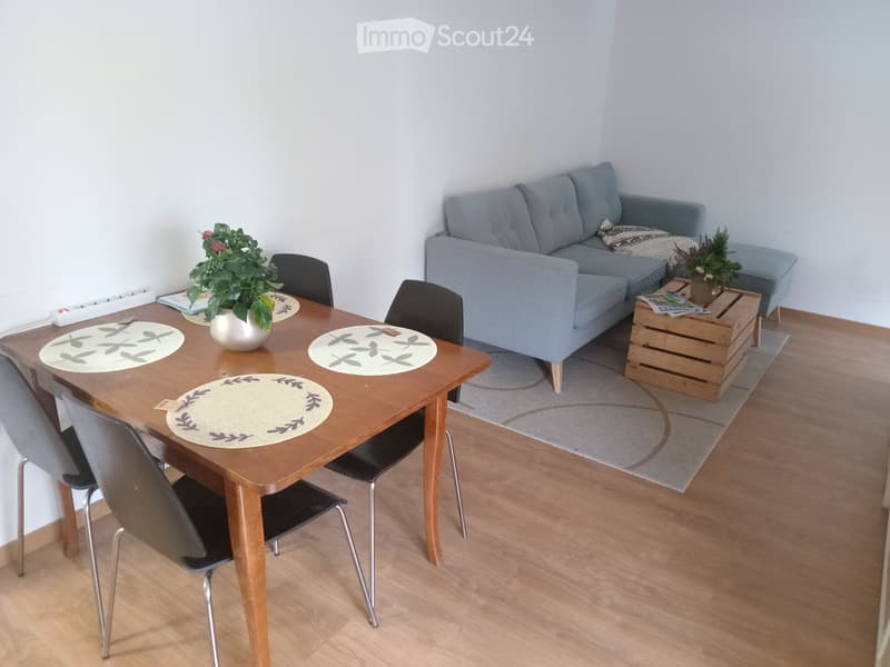 A modern living room with a grey sofa, wooden dining table, four chairs, a potted plant, and a wooden crate used as a side table.