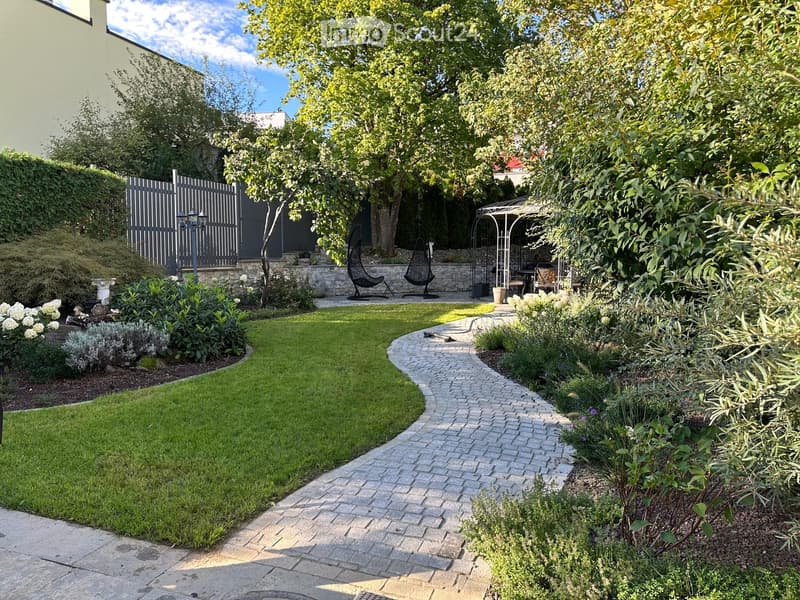 Well-maintained garden with a curved brick pathway, neatly trimmed lawn, various plants, trees, and outdoor seating area with hanging chairs and a small table.