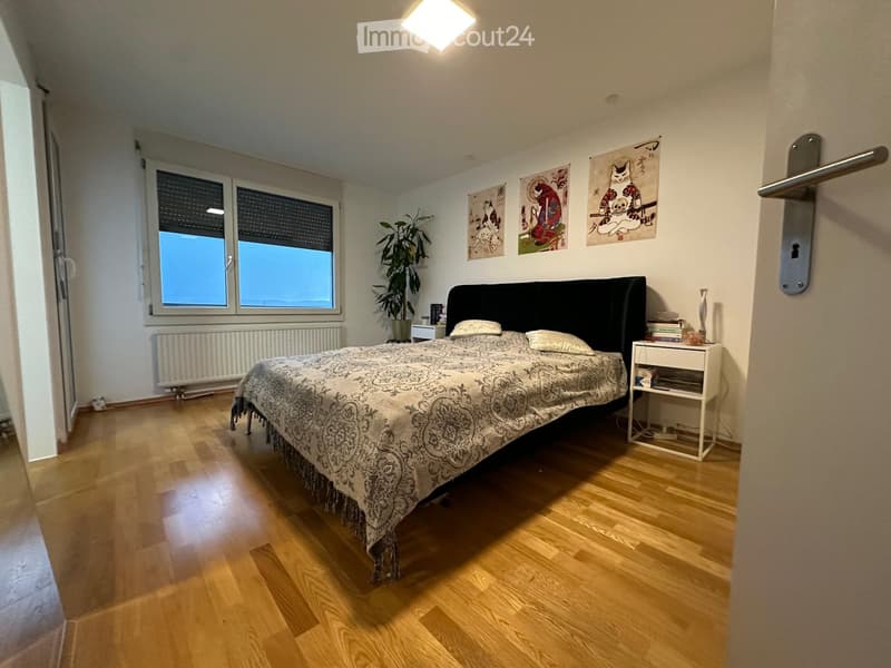 Spacious bedroom with wooden flooring, large window, bed with patterned bedspread, nightstand, plant, and artwork on walls.