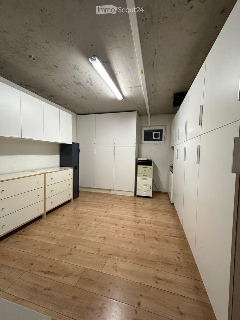 Empty room with light wooden flooring, white cabinetry, and a microwave mounted on the wall. The ceiling is concrete with exposed beams and a single fluorescent light fixture.