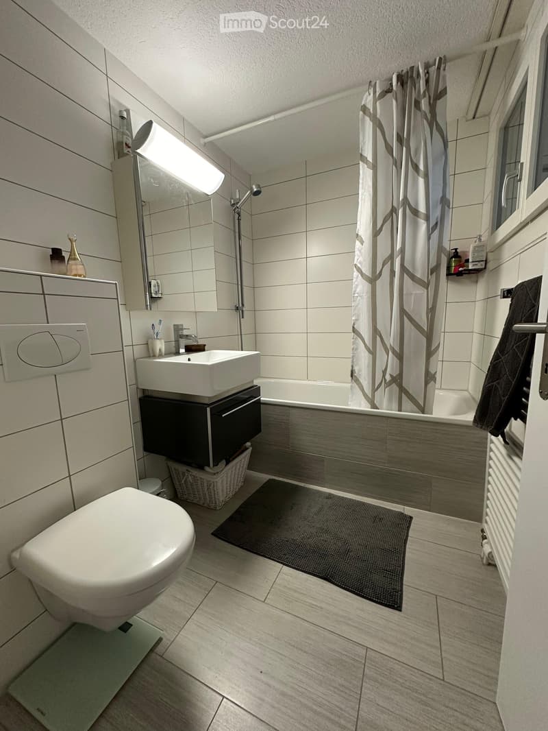 Modern bathroom with white tiles, a toilet, sink, bathtub, shower curtain, and a wicker basket on the floor.