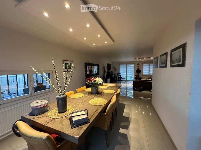 Dining area with a long wooden table, chairs, vase with flowers, placemats, and a view of a living room area with a sofa and mirror.