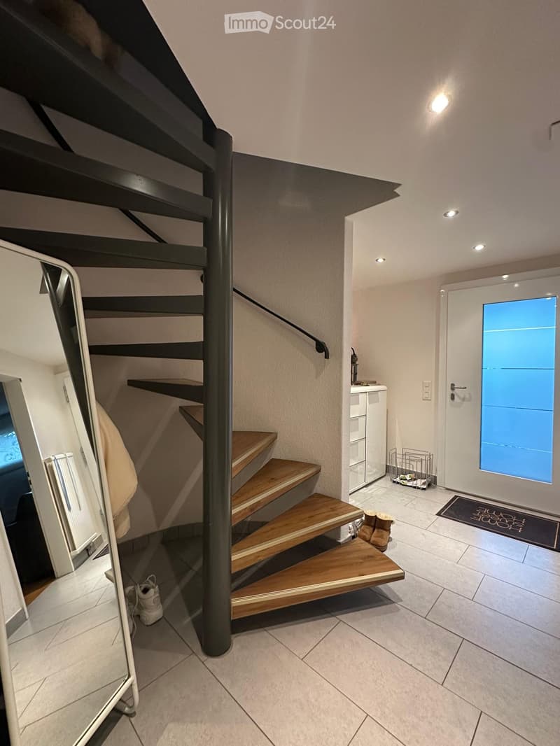 Modern hallway with a wooden staircase, white walls, and a small entryway area with a white door and a small cabinet.