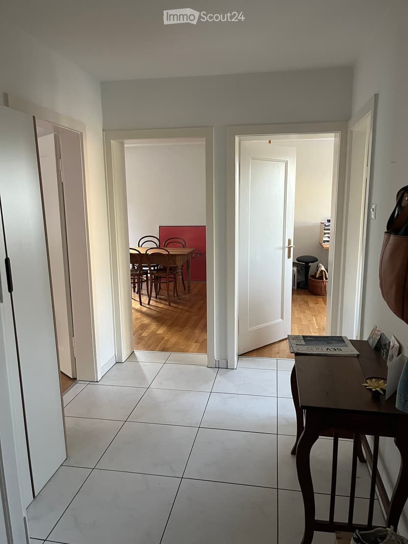 A clean, tiled hallway with white walls and multiple doors leading to different rooms, including a dining area and a room with a small table and chair.