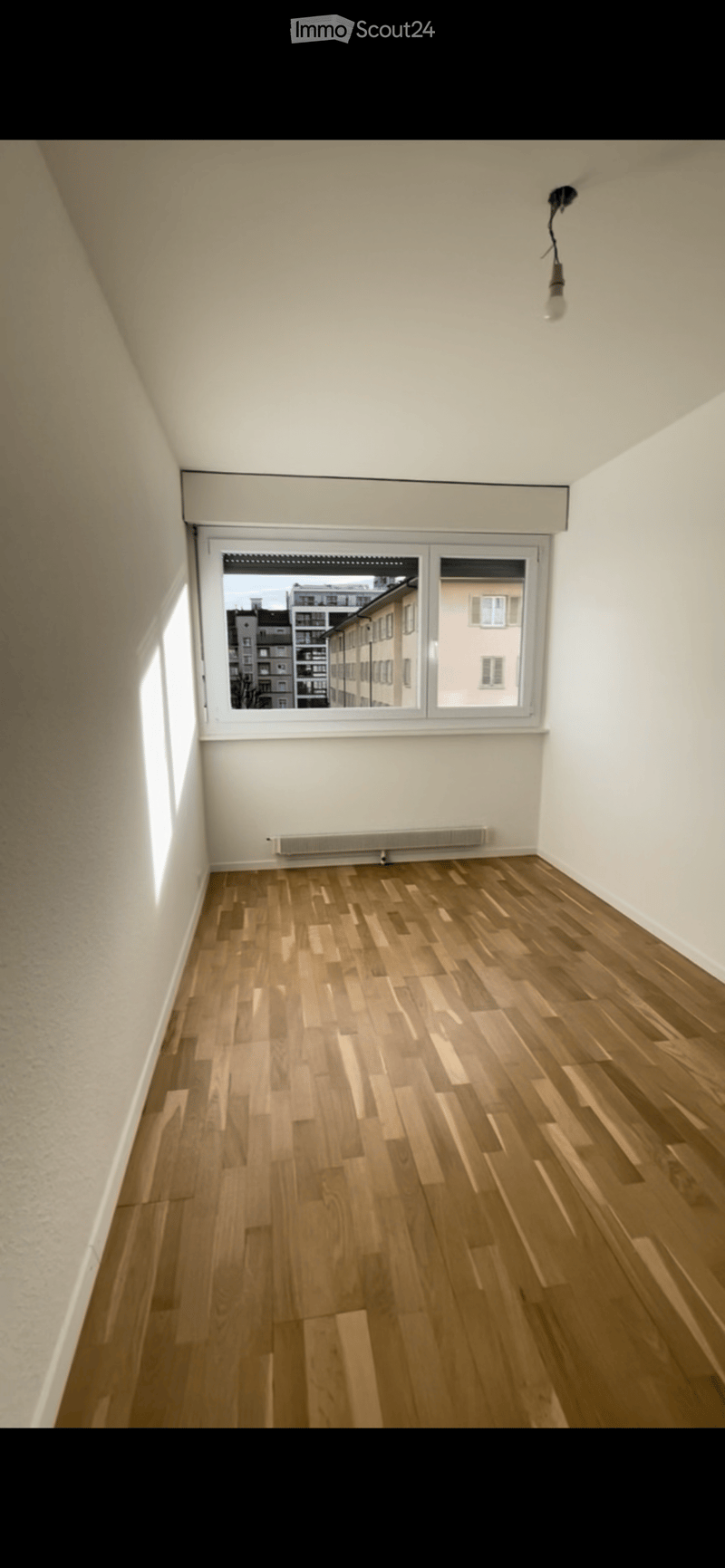 Empty room with wooden flooring, white walls, and a large window showing a cityscape view. No furniture or equipment present.