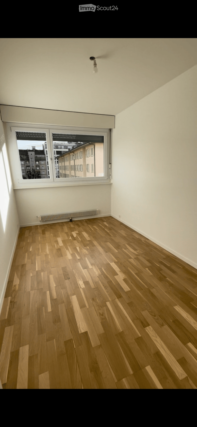 Empty room with wooden flooring, white walls, and large windows offering a view of the cityscape.