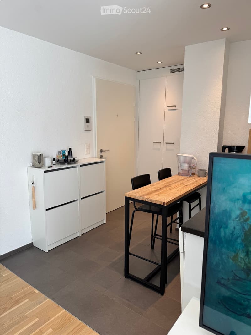 Modern kitchen with white cabinets, a wooden dining table, black chairs, and various appliances including a refrigerator and coffee maker.