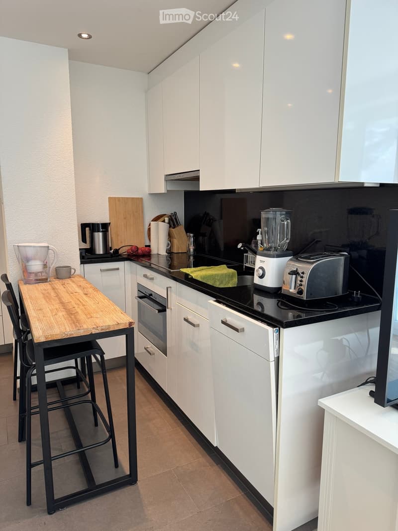 Modern kitchen with white cabinets, black countertop, wooden table, stools, and various appliances including a blender, toaster, and oven.