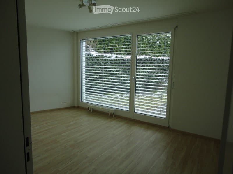 Empty room with wooden flooring, large window with blinds, and a small bench near the window.
