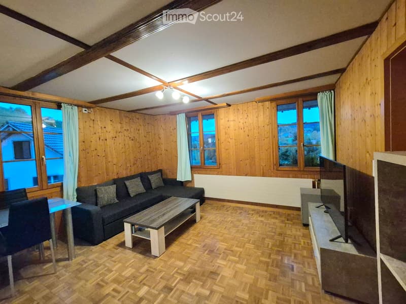 Living room with wooden walls, exposed wooden beams, hardwood floor, large windows, sofa, coffee table, dining table, chairs, and television.