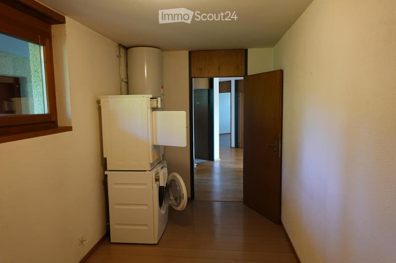 A narrow hallway with wooden flooring, white walls, and a window on the left side. A washing machine and water heater are stacked against the wall, and a door leads to another room.