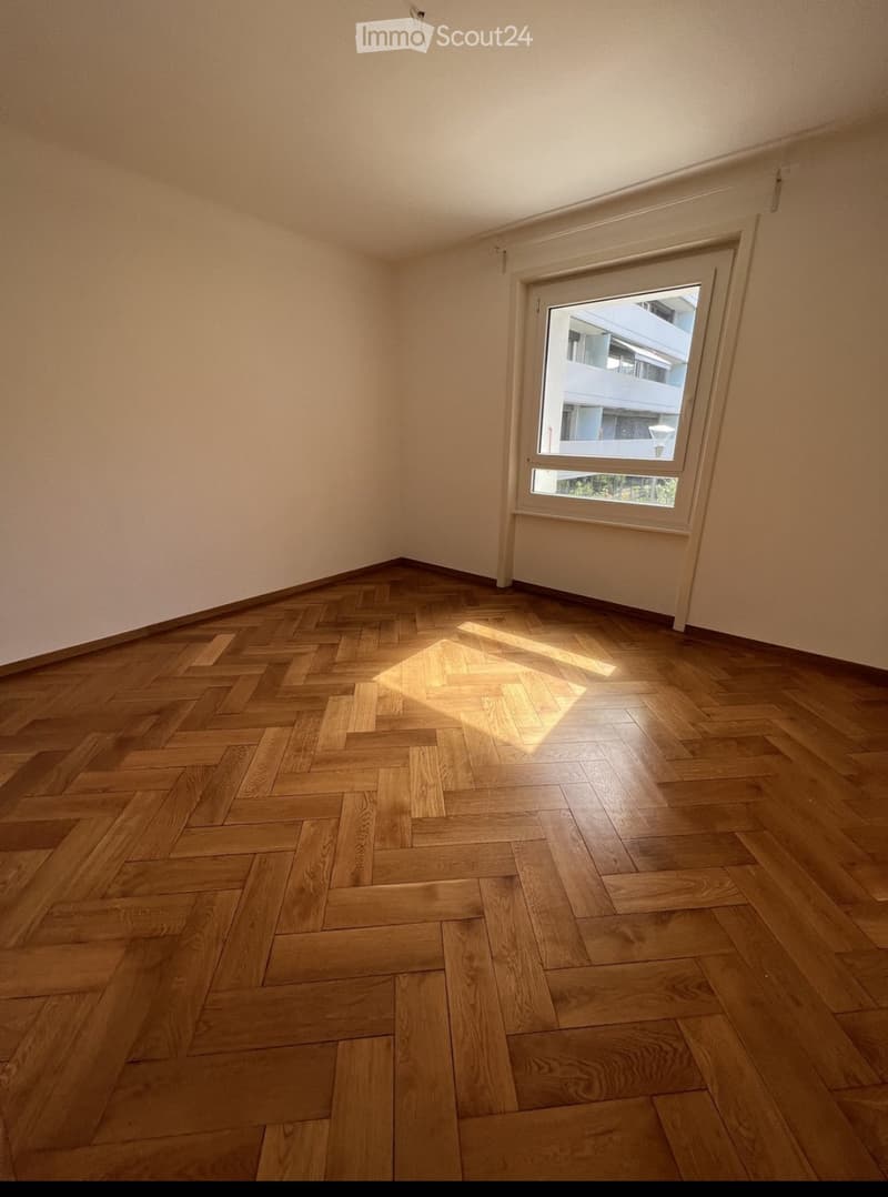 Empty room with wooden herringbone flooring and a single window showing a view of a building outside.