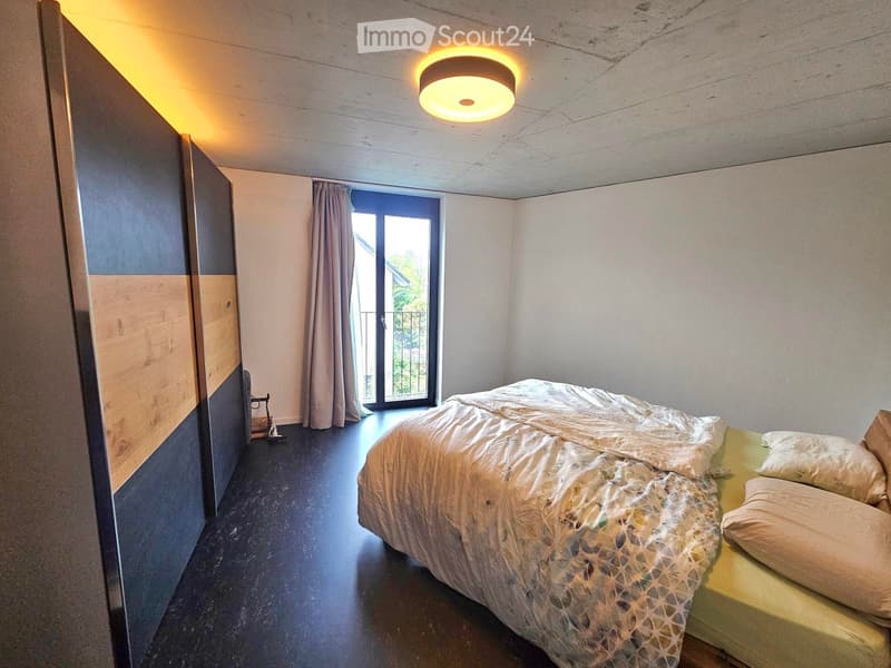 Modern bedroom with a bed, pillows, a wardrobe, a curtain, and a ceiling lamp. The room features a dark wooden floor and a large window with a view.