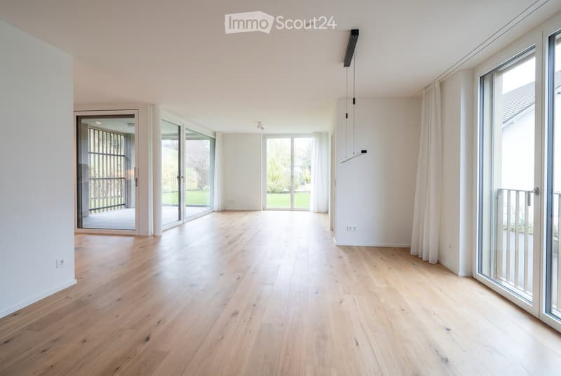 Bright, empty room with light wooden flooring, large glass sliding doors leading to a balcony, white walls, and modern lighting fixtures.