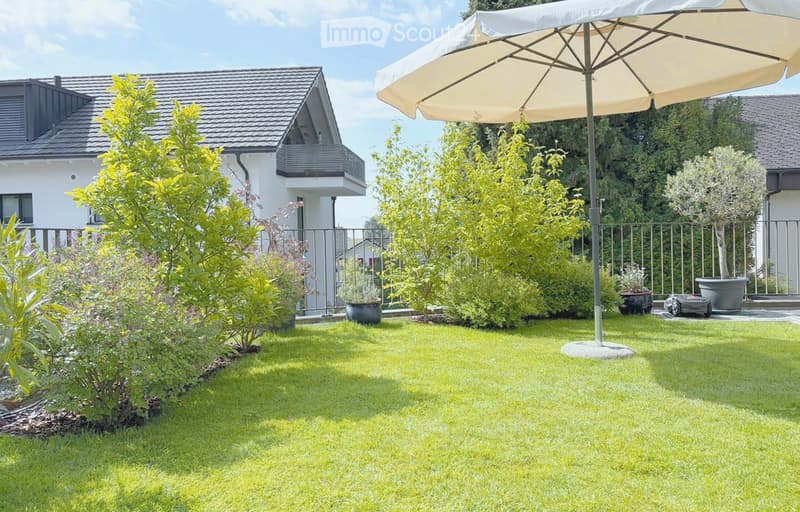 Well-maintained garden with green lawn, various shrubs and trees, a large patio umbrella, and a modern house in the background.