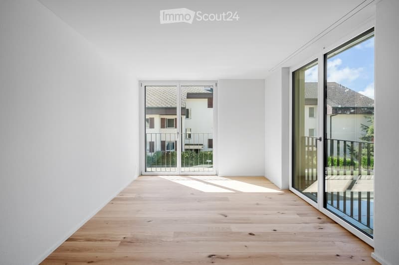 Empty room with light wooden flooring and white walls, featuring large glass doors leading to a balcony with a view of neighboring houses.