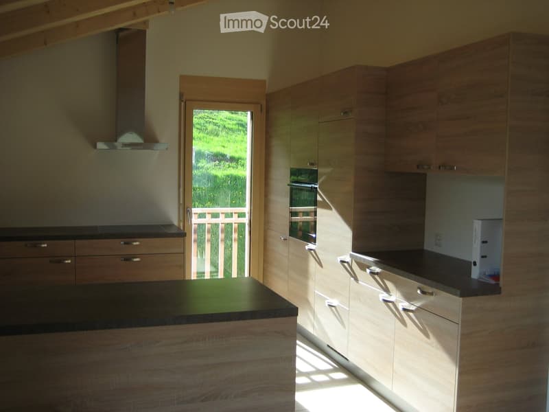 Modern kitchen with light wood cabinets, black countertops, and an island. Features an oven, range hood, and a door leading to a green outdoor area.